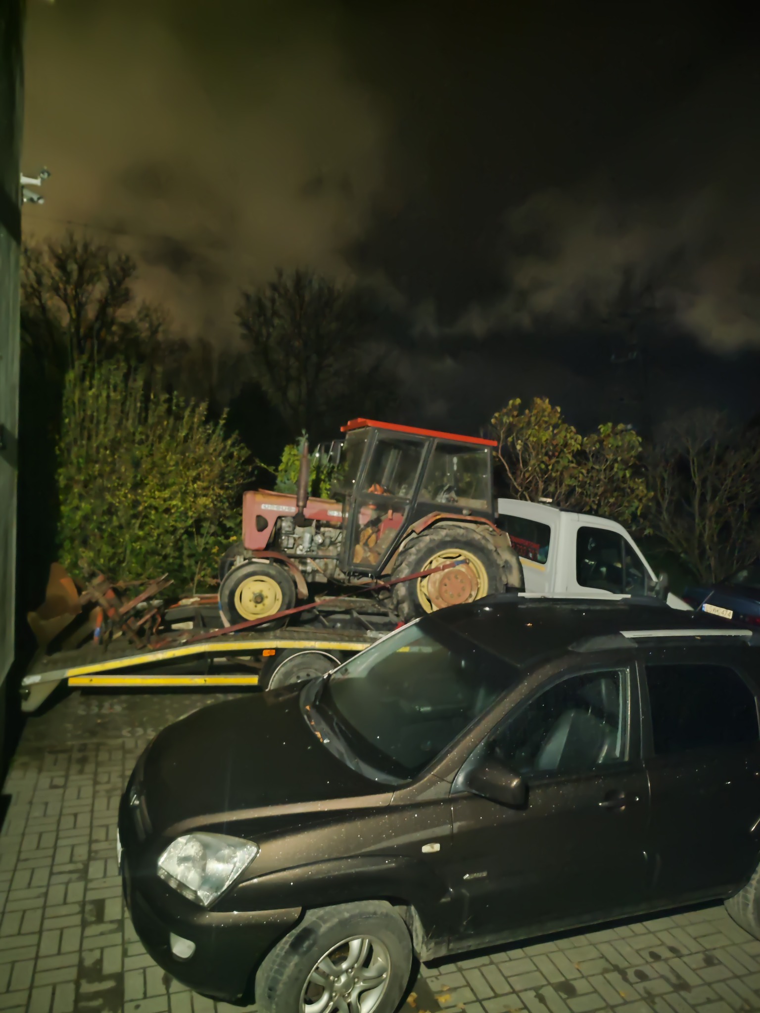 Nocne ujęcie lawety z czerwonym traktorem i osprzętem rolniczym, zaparkowanej obok ciemnego SUV-a na brukowanym podjeździe.