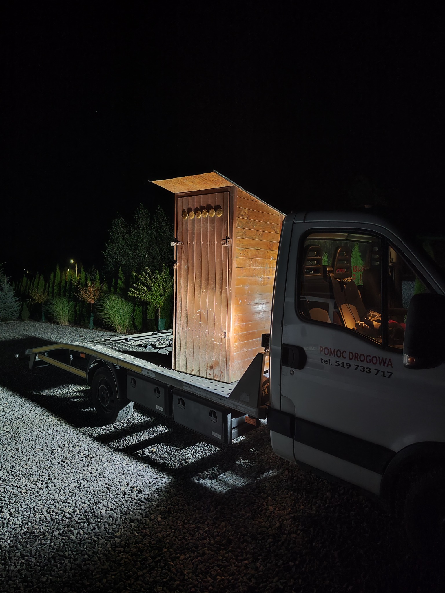 Nocny transport drewnianej budki na platformie lawety, widoczny numer telefonu 'Pomoc Drogowa' na pojeździe.