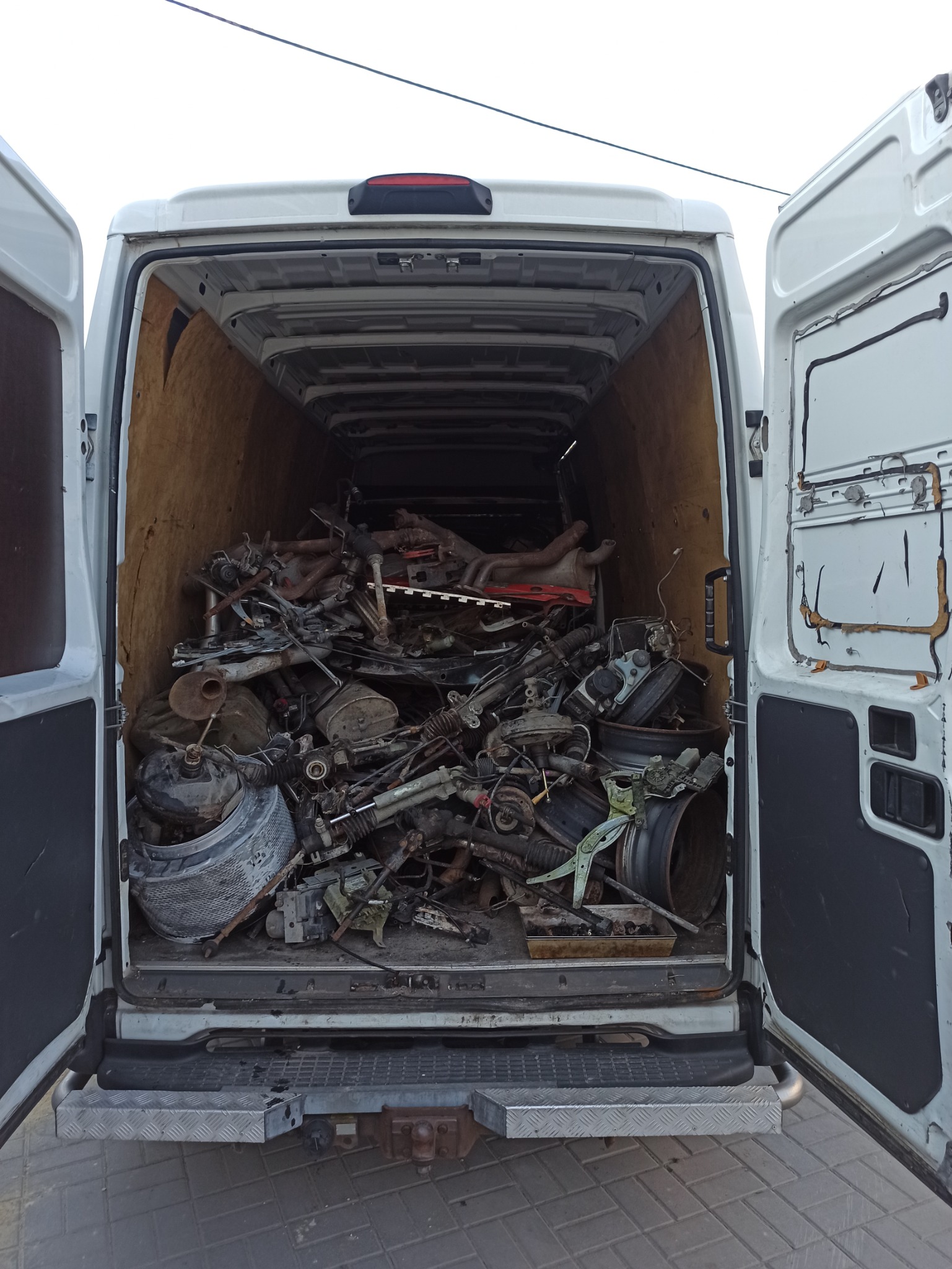 Wnętrze białego furgonetki z otwartymi drzwiami, wypełnione po brzegi różnorodnymi używanymi częściami samochodowymi, w tym kołami, amortyzatorami i elementami układu kierowniczego.
