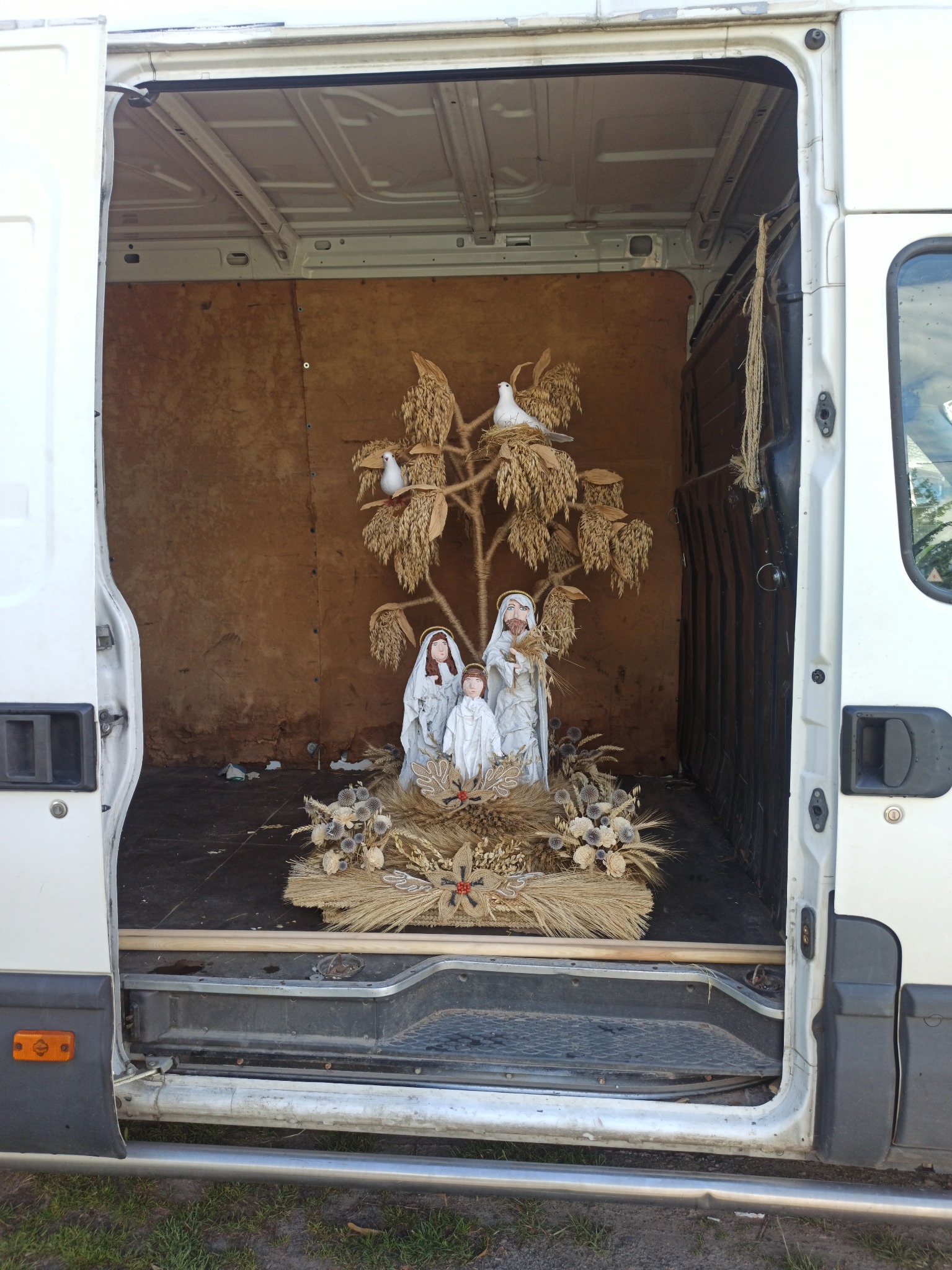 Wnętrze białego furgonetki z otwartymi drzwiami, w środku dekoracja religijna z figurkami Marii, Józefa i Jezusa, wykonana ze słomy i suszonych kwiatów, z dwoma białymi gołębiami na szczycie.