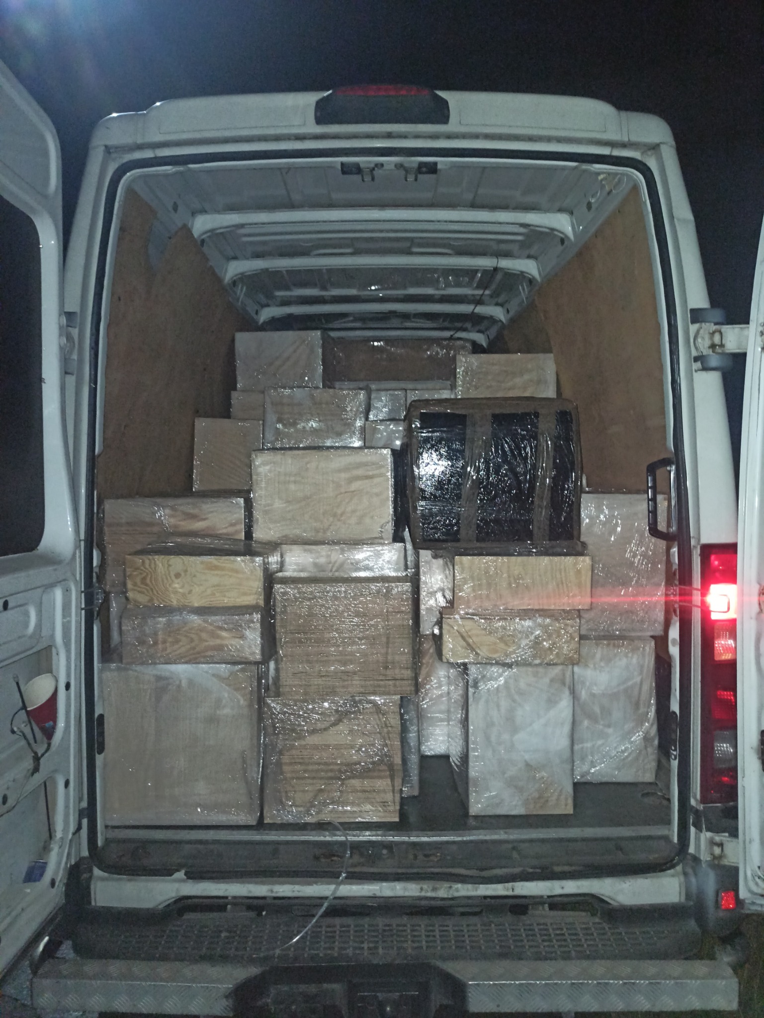 Wnętrze furgonetki wypełnionej po brzegi drewnianymi skrzyniami owiniętymi folią, widoczne otwarte tylne drzwi i kubek na drzwiach.