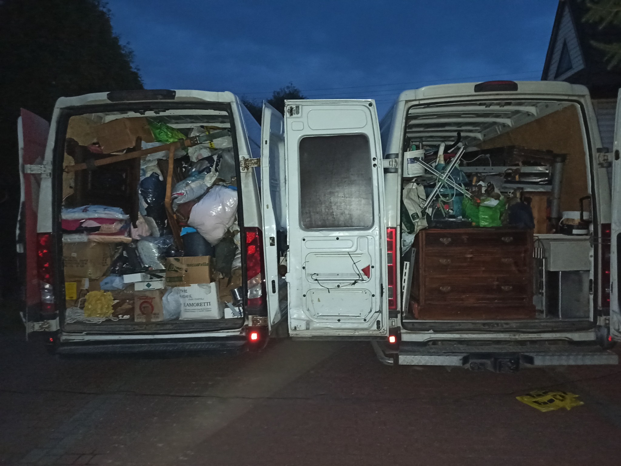 Dwa busy z otwartymi bagażnikami, wypełnione różnymi przedmiotami, w tym meblami, kartonami i workami, stoją obok siebie na zewnątrz, o zmierzchu.