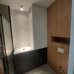 SkyProjects Bożena Wolińska - Nowoczesna łazienka z zabudową meblową: szafka umywalkowa w kolorze grafitowym, biała umywalka z okrągłym otworem, drewniana szafa z półką i gniazdkiem elektrycznym, szara podłoga z drobnym wzorem.