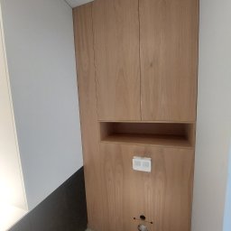 SkyProjects Bożena Wolińska - Drewniana szafa wnękowa z półką i panelem sterowania, widoczne otwory na rury, szare ściany i podłoga.