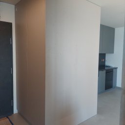 SkyProjects Bożena Wolińska - Wnętrze mieszkania w trakcie wykańczania, widoczna zabudowa meblowa w odcieniach szarości, fragment kuchni w tle oraz zabezpieczona podłoga.