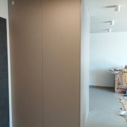 SkyProjects Bożena Wolińska - Zamknięta, beżowa szafa wnękowa z gładkimi drzwiami, widoczna w korytarzu podczas montażu, z kartonami i resztkami materiałów budowlanych na podłodze.