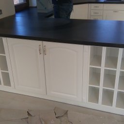 SkyProjects Bożena Wolińska - Wyspa kuchenna z białymi szafkami z frezowanymi drzwiczkami i czarnym blatem, widoczne otwarte półki z kratownicą i fragment podłogi w trakcie remontu.