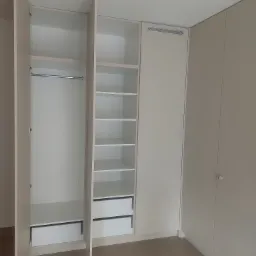 Wnętrze szafy wnękowej z otwartymi drzwiami, widoczne p&oacute;łki, szuflady i drążek na ubrania, minimalistyczny design w jasnych odcieniach.