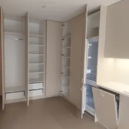 Beżowa zabudowa meblowa z otwartymi szafami, półkami i lodówką w minimalistycznym wnętrzu z widocznymi przewodami elektrycznymi na suficie.