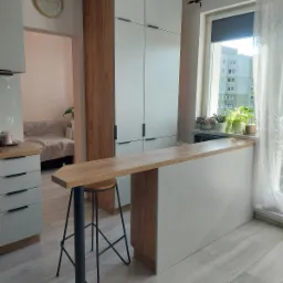 Wnętrze kuchni z drewnianym blatem barowym, wysokim krzesłem na tle białych szafek i okna z roślinami.