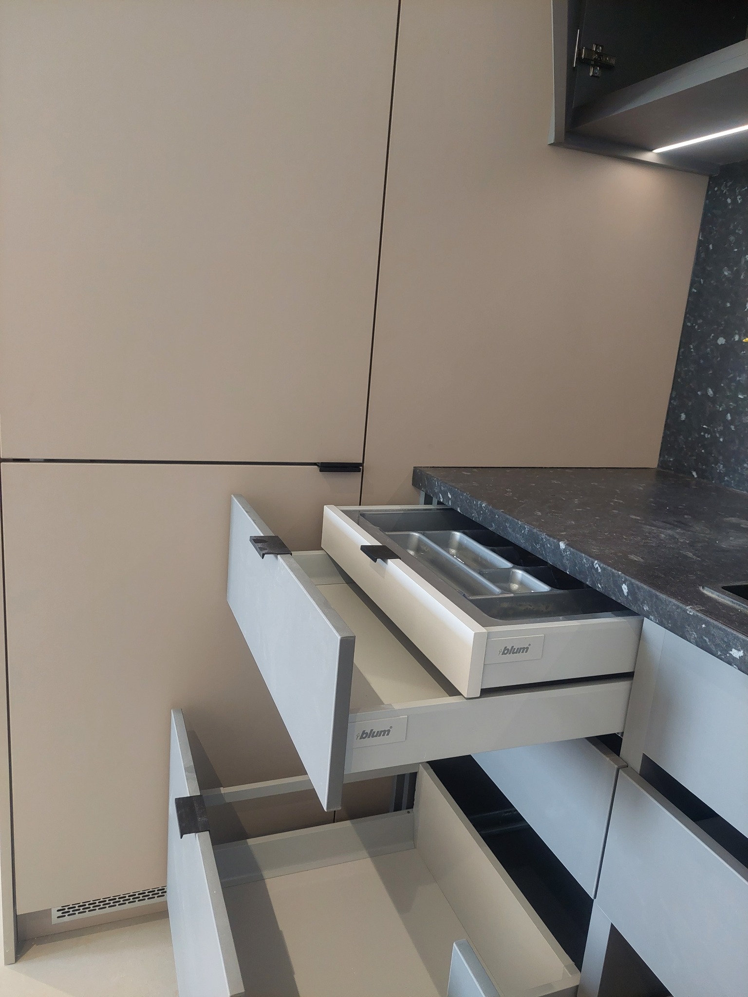 Szare szuflady kuchenne z organizerem na sztućce i systemem Blum, widoczne z bliska, z czarnym blatem i beżowymi frontami szafek.