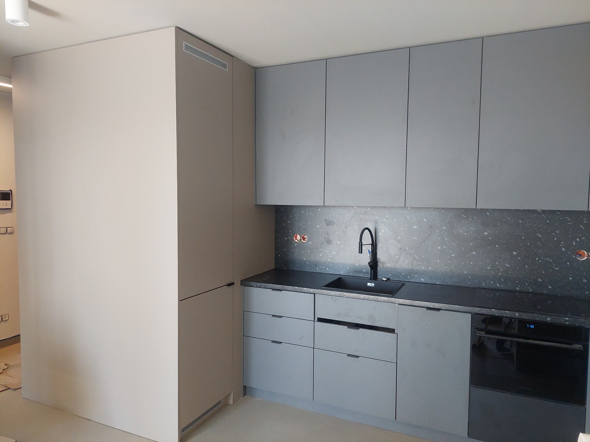 Szare matowe meble kuchenne z czarnym blatem i zlewozmywakiem, widoczny piekarnik i fragment ściany z szarą mozaiką.
