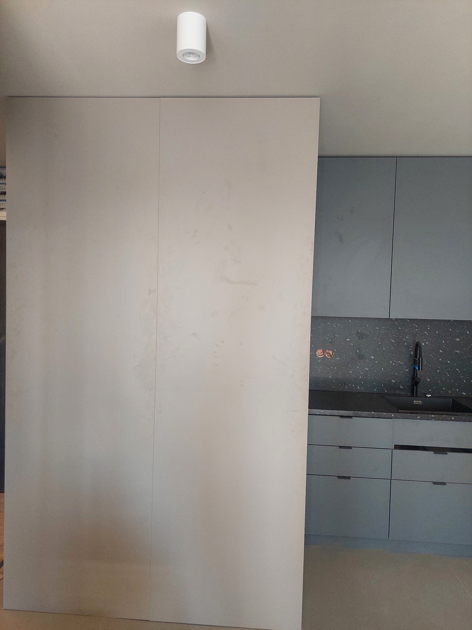 Minimalistyczna kuchnia z szarymi szafkami i blatem w ciemne plamki, widziana zza jasnej, przesuwnej ściany, oświetlona przez okrągłą lampę sufitową.