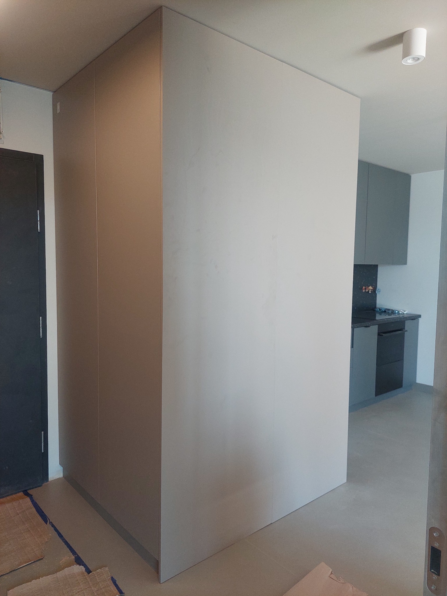 Wnętrze mieszkania w trakcie wykańczania, widoczna zabudowa meblowa w odcieniach szarości, fragment kuchni w tle oraz zabezpieczona podłoga.