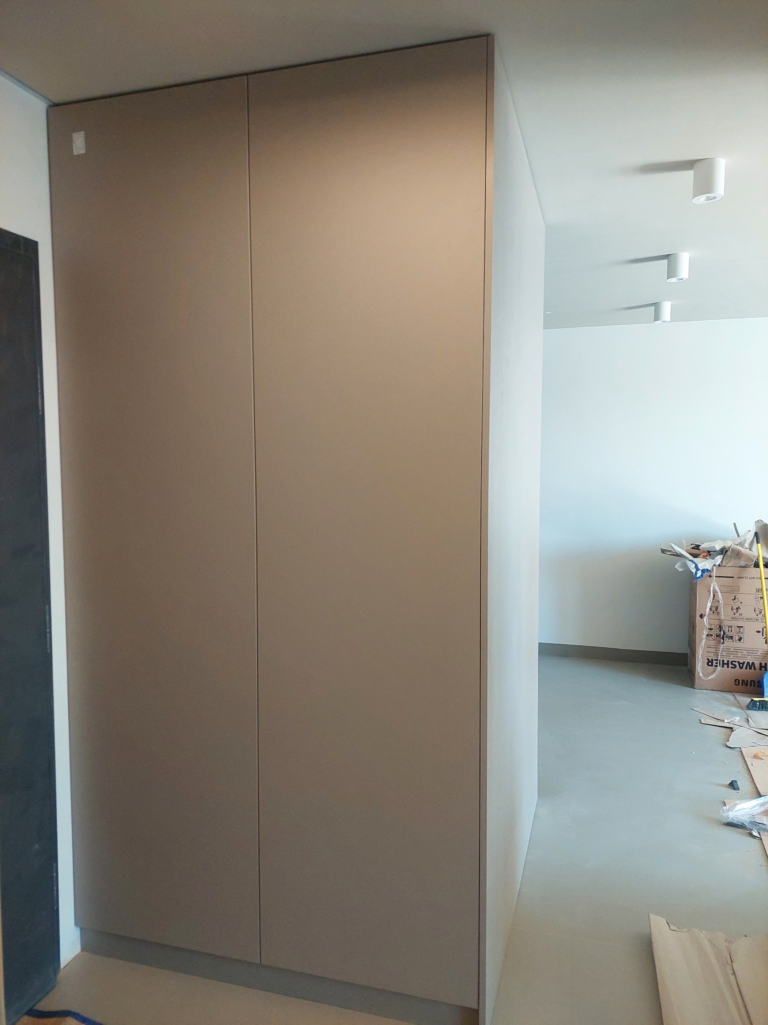 Zamknięta, beżowa szafa wnękowa z gładkimi drzwiami, widoczna w korytarzu podczas montażu, z kartonami i resztkami materiałów budowlanych na podłodze.
