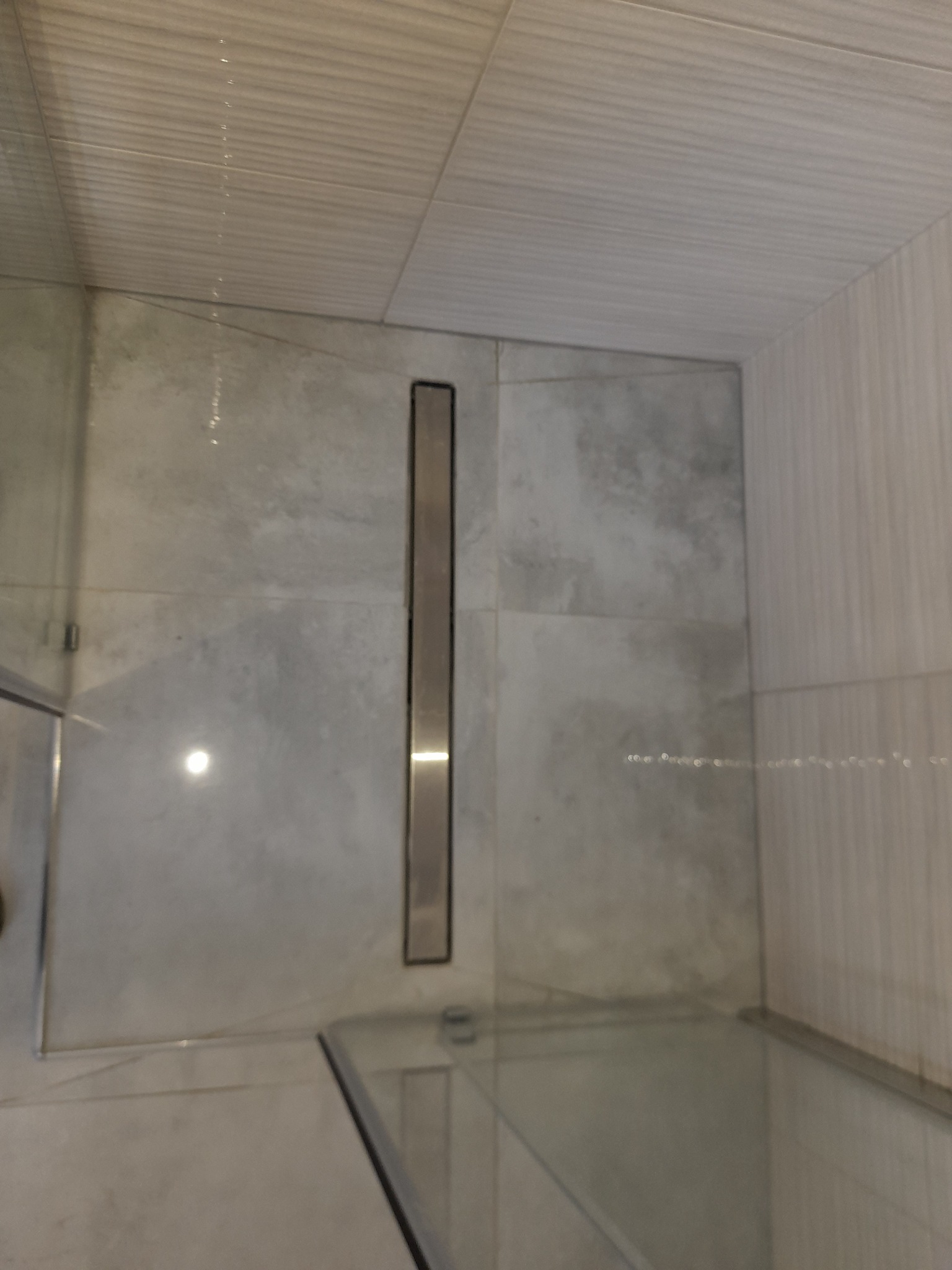 Szczegółowe ujęcie odpływu liniowego w kabinie prysznicowej, z widocznymi szarymi płytkami na ścianach i podłodze oraz fragmentem szklanej kabiny.