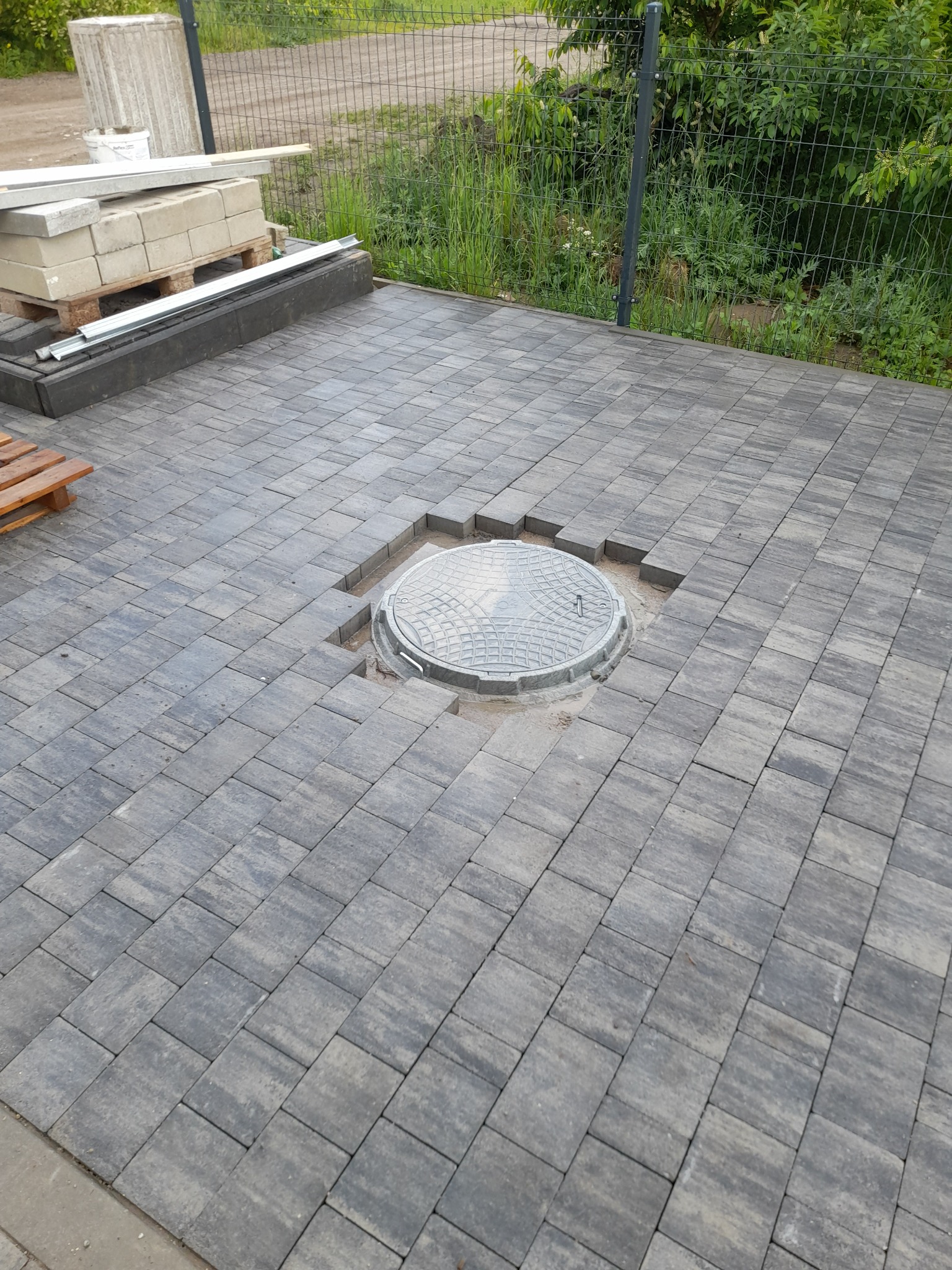 Układanie szarej kostki brukowej wokół studzienki kanalizacyjnej na zewnątrz, widoczne niedokończone krawędzie i zapas materiału budowlanego w tle.