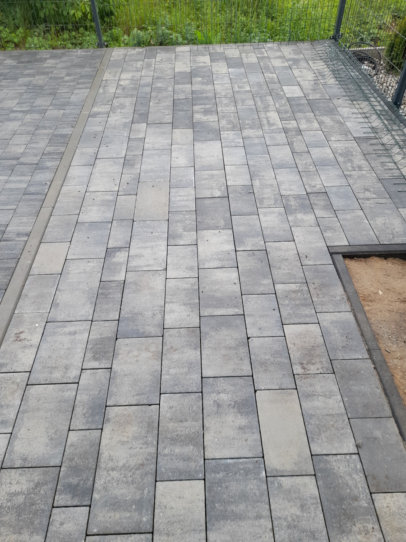 Ułożona szara kostka brukowa na tarasie, widoczna krawędź z piaskiem i ogrodzenie z siatki w tle.