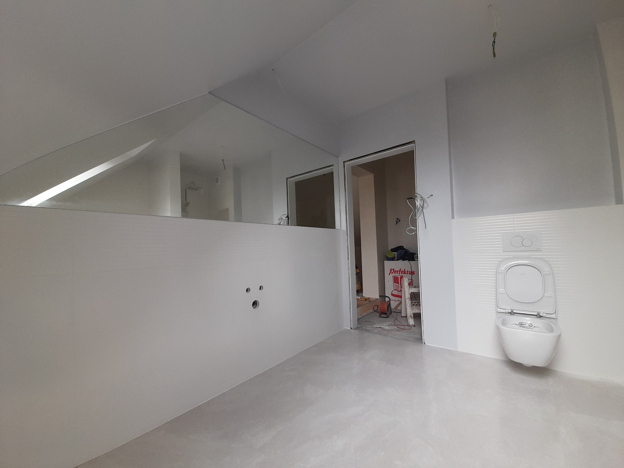 Półka z szybą w łazience ze świeżo położonymi białymi płytkami, widoczny podwieszany sedes i otwór drzwiowy prowadzący do kolejnego pomieszczenia z narzędziami.