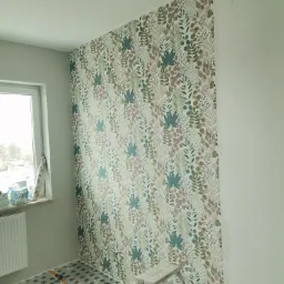 Ściana częściowo pokryta tapetą w roślinny wzór, widoczna drabina, folia ochronna na podłodze, rolka do tapetowania i akcesoria, fragment okna z rolką tapety.