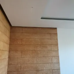 Ściana wykończona panelami imitującymi drewno, sufit podwieszany z oświetleniem punktowym, widoczne łączenie paneli na ścianie.