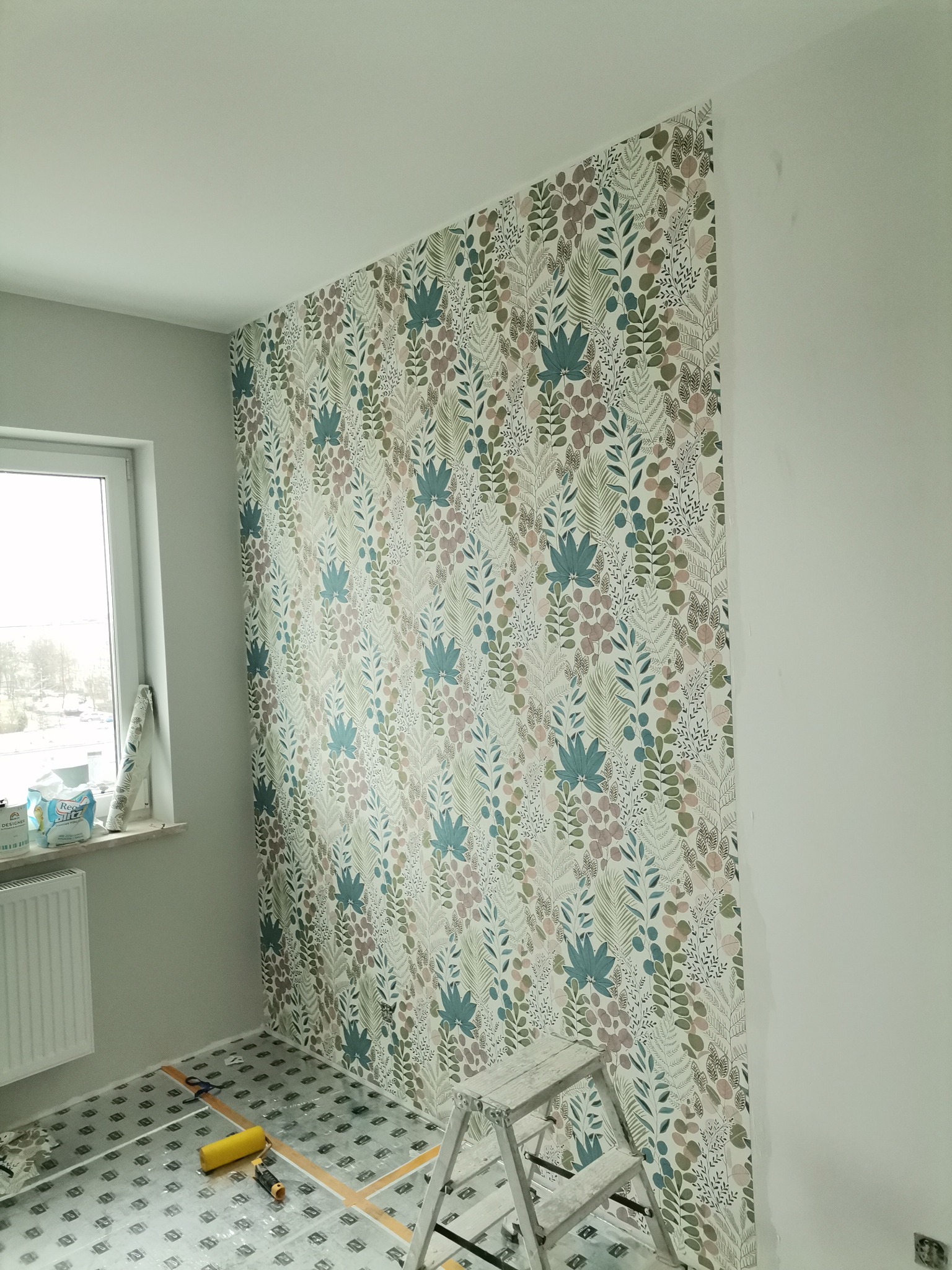 Ściana częściowo pokryta tapetą w roślinny wzór, widoczna drabina, folia ochronna na podłodze, rolka do tapetowania i akcesoria, fragment okna z rolką tapety.