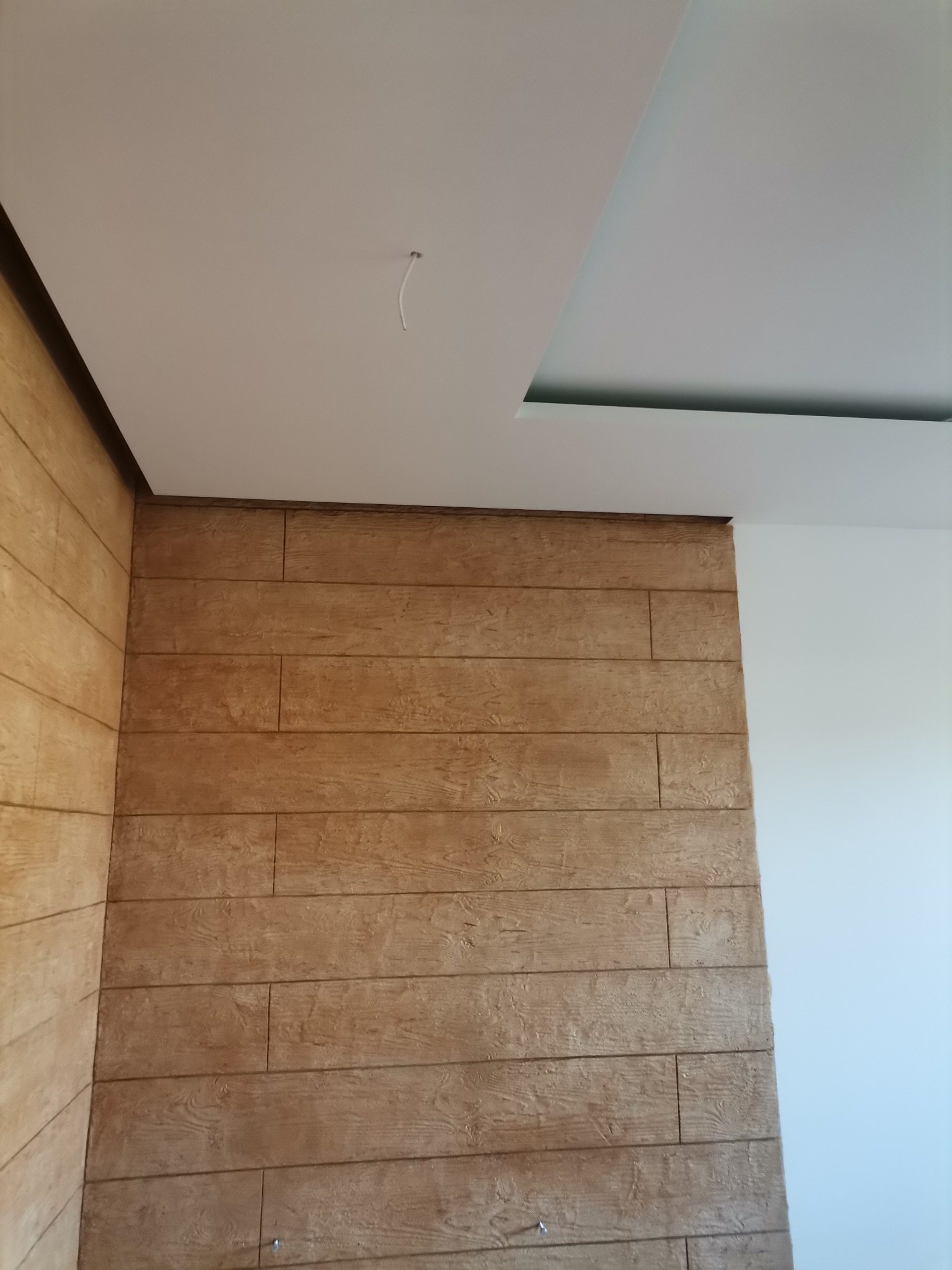 Ściana wykończona panelami imitującymi drewno, sufit podwieszany z oświetleniem punktowym, widoczne łączenie paneli na ścianie.