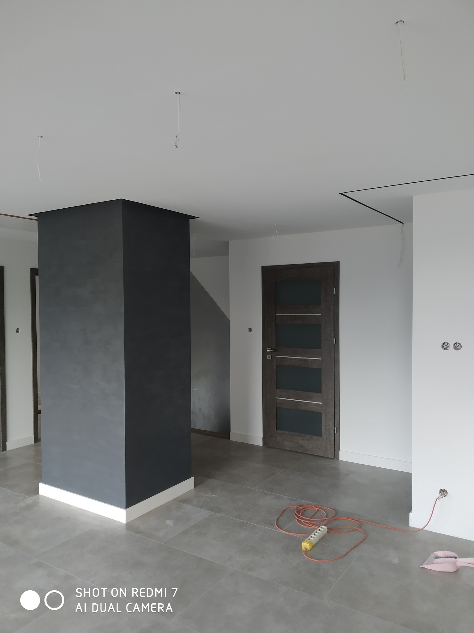 Wnętrze w trakcie prac wykończeniowych: pomalowany na szaro filar, szara podłoga, zamontowane drzwi wewnętrzne, widoczne kable i przedłużacz na podłodze.