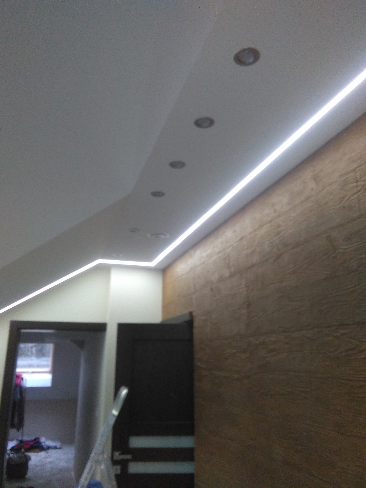 Nowoczesne oświetlenie LED liniowe pod sufitem podwieszanym, z widocznymi punktami świetlnymi, ściana wykończona panelami imitującymi drewno, otwarte drzwi do innego pomieszczenia.