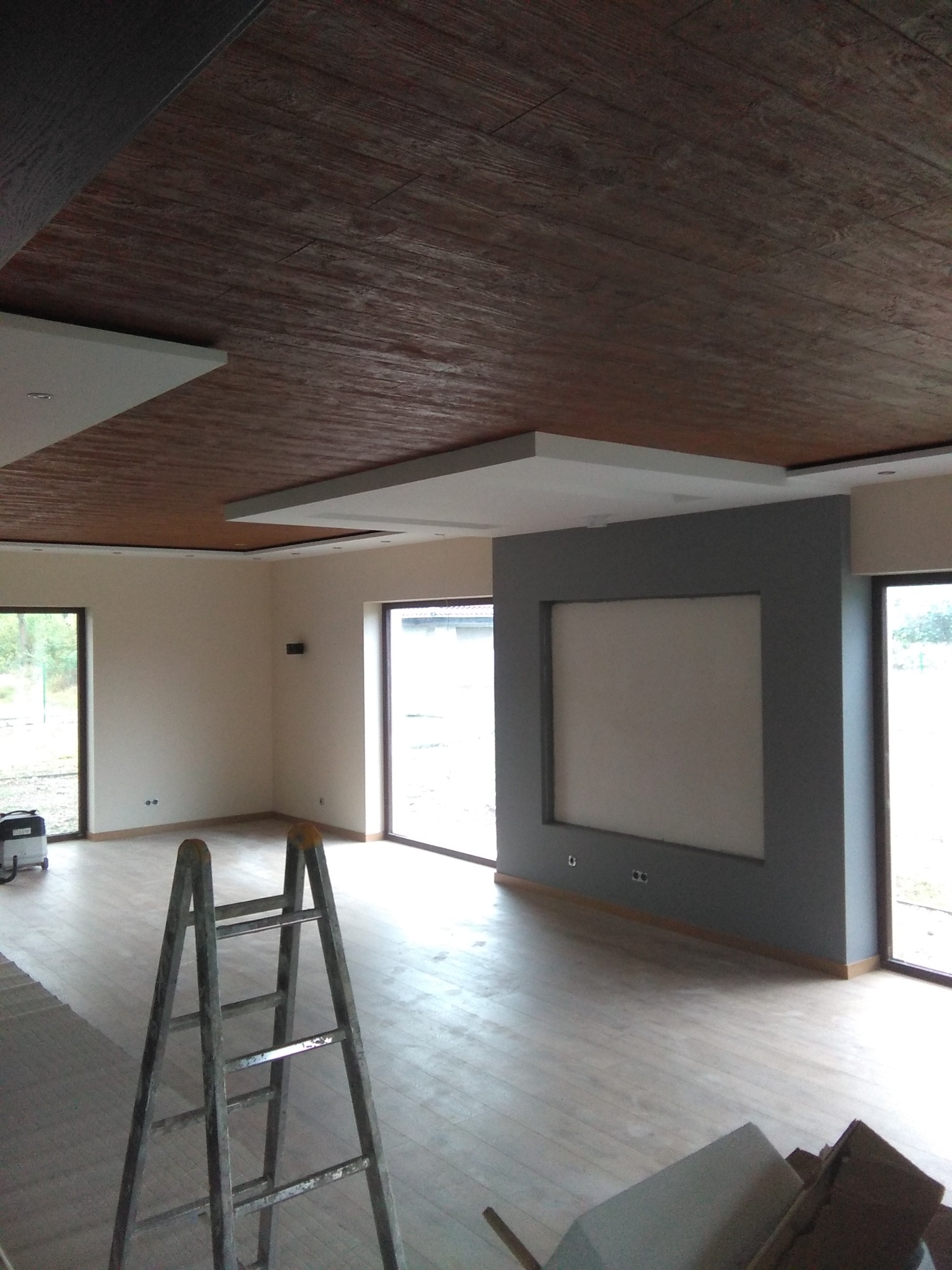 Wnętrze w trakcie prac wykończeniowych, widoczny drewnopodobny sufit z elementami podwieszanymi, szara ściana z wnęką, drabina oraz odkurzacz przemysłowy.