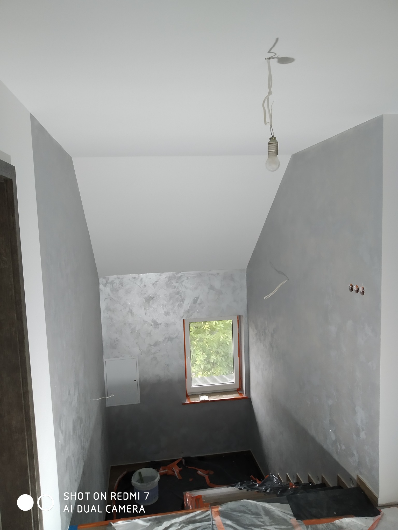 Wnętrze klatki schodowej w trakcie prac wykończeniowych, ściany z efektem dekoracyjnym, zabezpieczone schody, surowa żarówka zwisająca z sufitu.