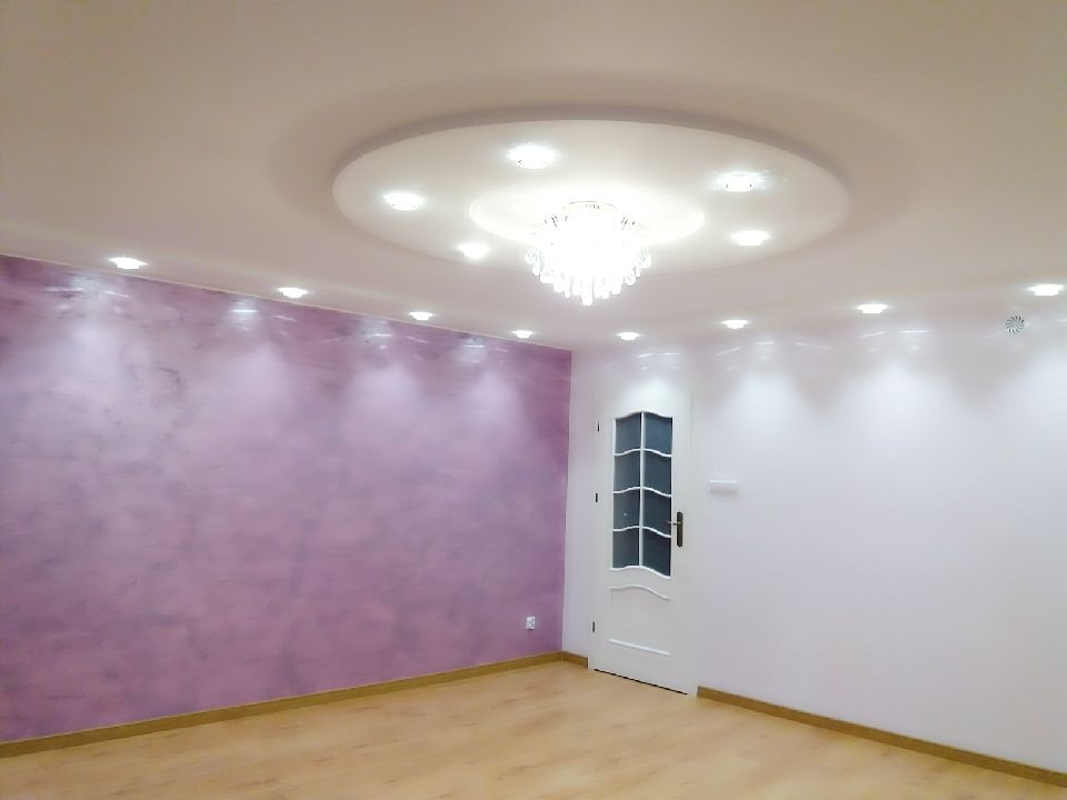 Wykończone wnętrze: ściana w odcieniach fioletu z efektem dekoracyjnym, drewniana podłoga, białe ściany z halogenami i ozdobny sufit z żyrandolem, białe drzwi z szybami.