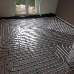 Białe rury ogrzewania podłogowego ułożone spiralnie na folii z czerwoną siatką, z widocznym oknem i wiadrem w tle, w trakcie instalacji.