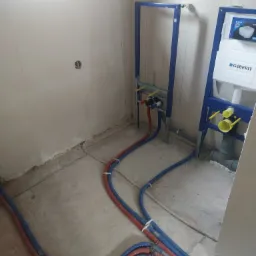 Widok na surową łazienkę w trakcie prac hydraulicznych: nieotynkowane ściany, rury w izolacji czerwono-niebieskiej prowadzone po podłodze, stelaż podtynkowy WC marki Geberit w niebieskiej ramie.