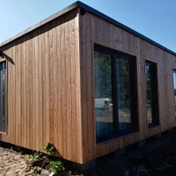 Zewnętrzny widok z boku na nowoczesny dom modułowy z drewnianą elewacją, czarnymi ramami okiennymi i drzwiami, na betonowych fundamentach, z widocznym samochodem dostawczym w tle.