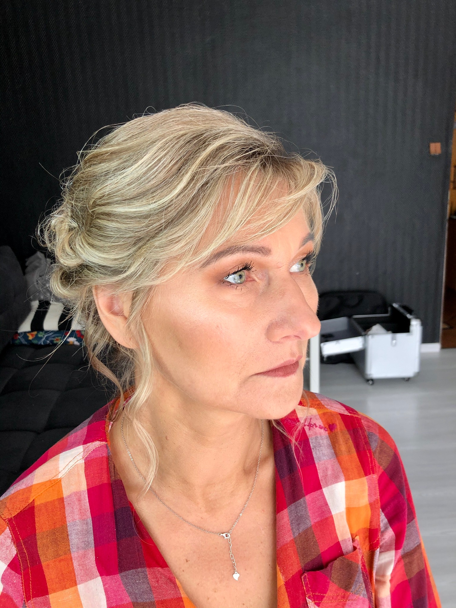 Profil kobiety z upiętymi blond włosami i delikatnym brązowym cieniem na powiekach, ubrana w koszulę w czerwoną kratę, z widoczną srebrną zawieszką na szyi, w tle czarny dywan i biała walizka...