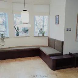 Narożna zabudowa siedziska z tapicerowanymi poduchami w odcieniach brązu, z dodatkowymi stolikami, na tle parkietu i okien z roślinami.