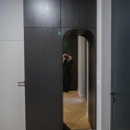 AWENAmeble - Ciemna zabudowa szafy z lustrem w kształcie łuku, odbijającym wnętrze mieszkania z parkietem w jodełkę, widoczny fotograf robiący zdjęcie.