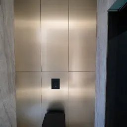 Nowoczesna toaleta podwieszana w zabudowie z paneli w odcieniach złota, z czarnym przyciskiem spłukującym i marmurową okładziną ściany.