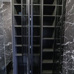 AWENAmeble - Czarna szafa wnękowa z przeszklonymi drzwiami harmonijkowymi na tle ściany z czarnego marmuru, widoczny fragment toalety.