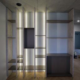 AWENAmeble - Nowoczesna zabudowa regałowa z podświetleniem LED, szare półki i szafka na tle betonowego sufitu i parkietu w jodełkę.