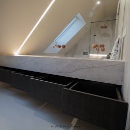 AWENAmeble - Łazienka na poddaszu z marmurową umywalką i otwartymi szufladami z ciemnego drewna; widoczne miedziane baterie i kabina prysznicowa w tle.