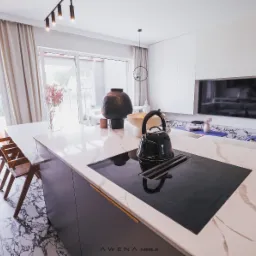 Nowoczesna kuchnia z marmurowym blatem, czarną płytą indukcyjną i czajnikiem. W tle salon z telewizorem i wyjściem na taras.