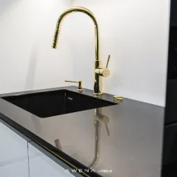 Czarny zlew wpuszczony w szary blat, bateria i dozownik w kolorze złotym, białe szafki w tle, nowoczesny design.