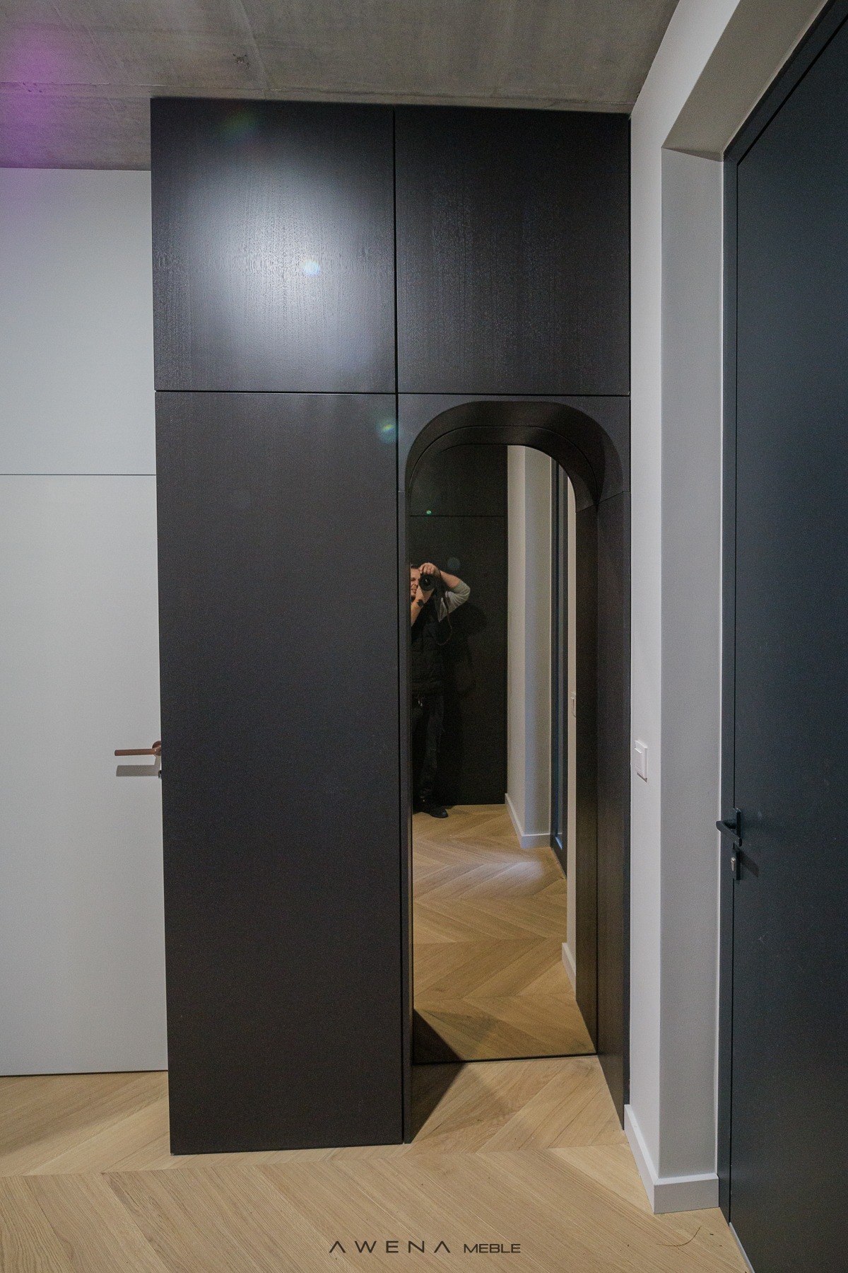 Ciemna zabudowa szafy z lustrem w kształcie łuku, odbijającym wnętrze mieszkania z parkietem w jodełkę, widoczny fotograf robiący zdjęcie.