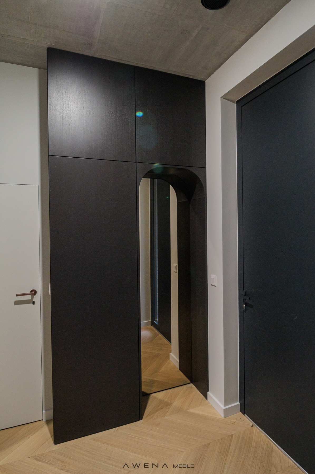 Nowoczesna, czarna szafa wnękowa z lustrem w kształcie łuku, wkomponowana w przedpokój z parkietem w jodełkę i surowym betonowym sufitem.