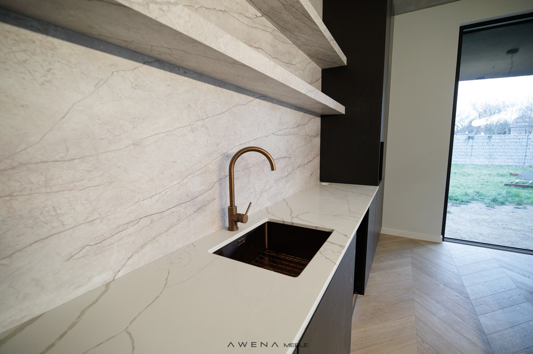Nowoczesna kuchnia z białym blatem imitującym marmur, miedzianą armaturą i otwartymi półkami. Widoczna czarna, minimalistyczna zabudowa i podłoga ułożona w jodełkę. Duże okno z widokiem na ogród.