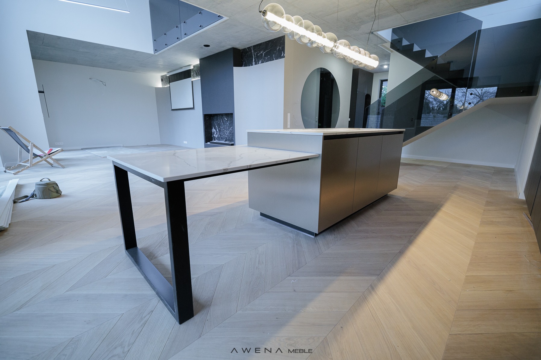 Nowoczesna wyspa kuchenna z blatem z imitacji marmuru, czarnymi nogami i szarymi szafkami, na parkiecie w jodełkę, w surowym wnętrzu z betonowym sufitem i szklaną balustradą schodów.