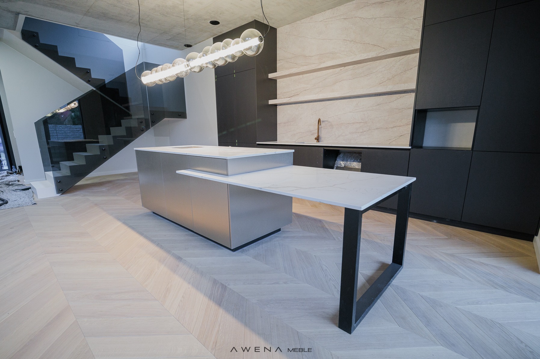 Nowoczesna kuchnia z wyspą i blatem z białego marmuru, czarne szafki, podłoga z jasnego drewna ułożona w jodełkę, przeszklone schody w tle.