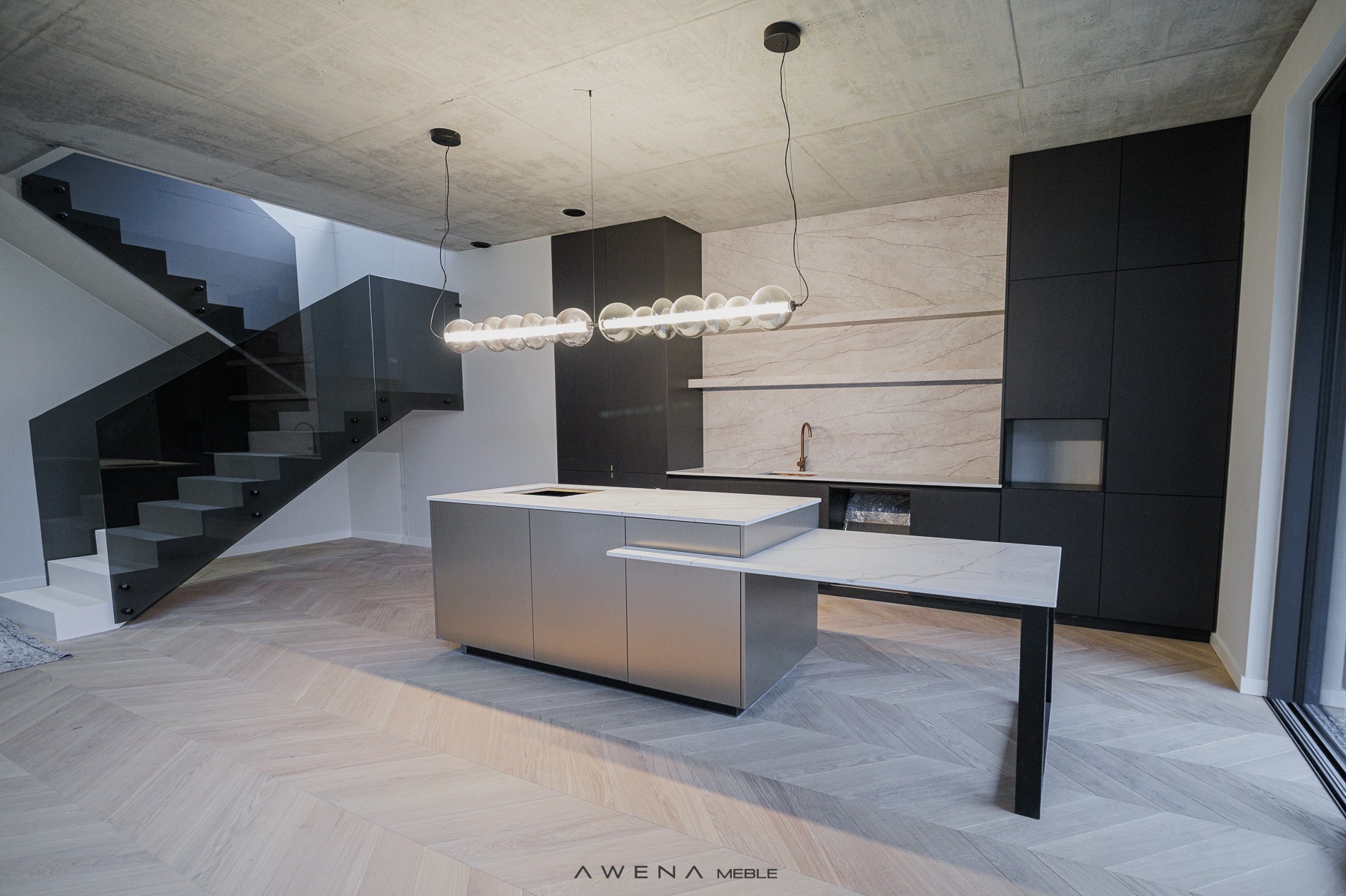 Nowoczesna kuchnia z wyspą w odcieniach szarości i czerni, oświetlona designerską lampą z kulistymi kloszami, parkiet ułożony we wzór jodełki, widoczne schody ze szklaną balustradą i surowy...